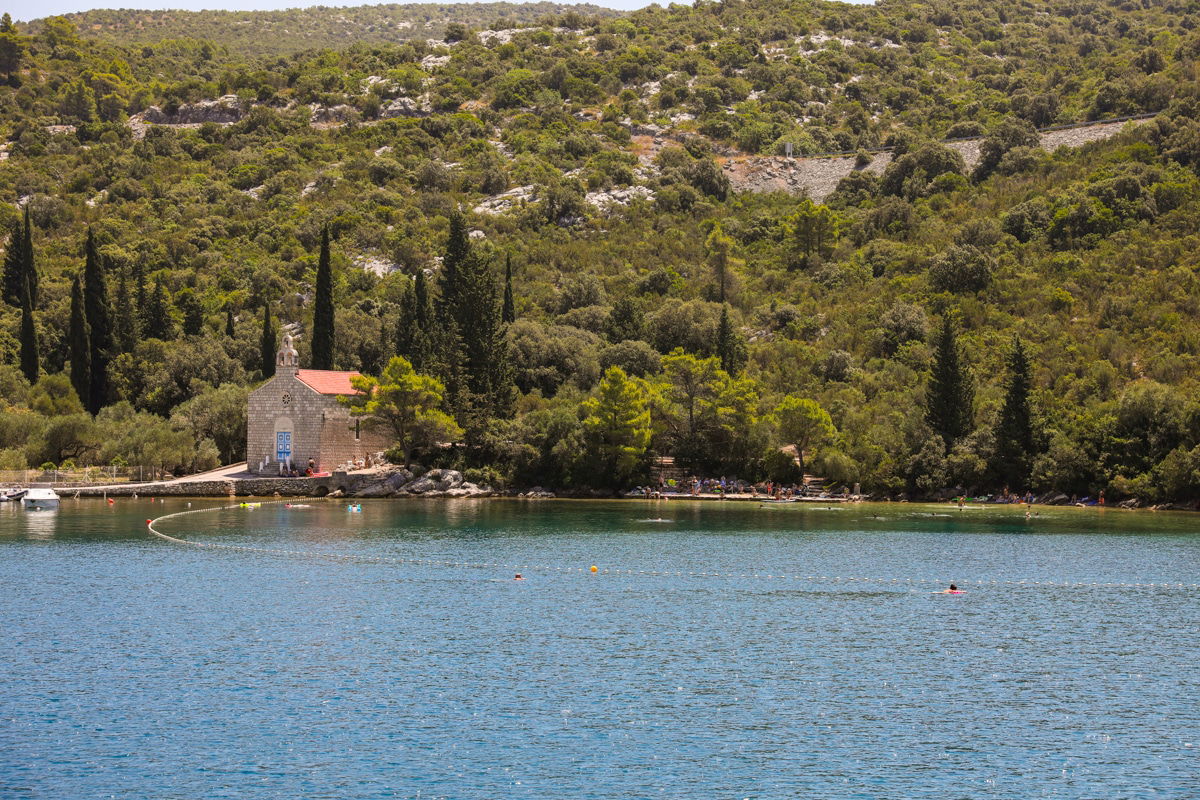 A small stone building with a red roof is situated near the shore of a calm, forested Slano Bay. The surrounding area is densely covered with trees, reminiscent of the serene landscapes by The Adriatic Sea.