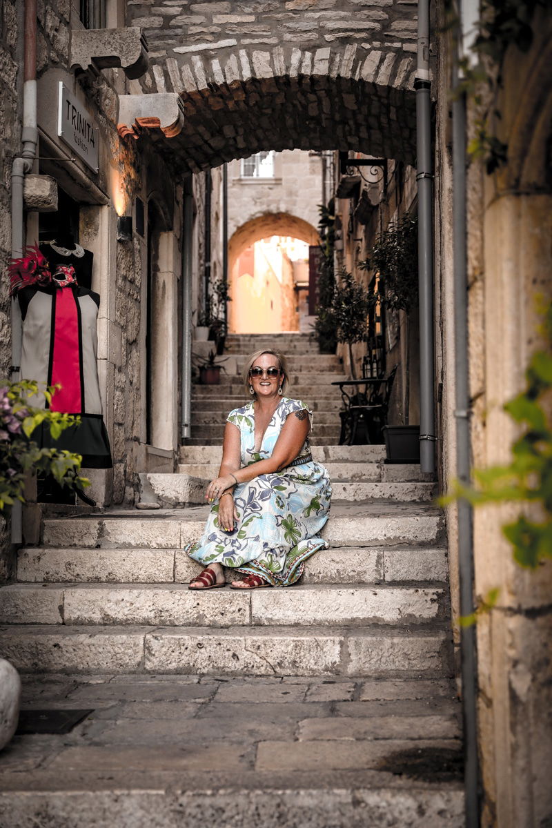 SJ loral dress sits on stone steps in a narrow, picturesque alley with archways and rustic buildings, reminiscent of the charming streets you might explore during a luxury cruise stop in Dubrovnik.